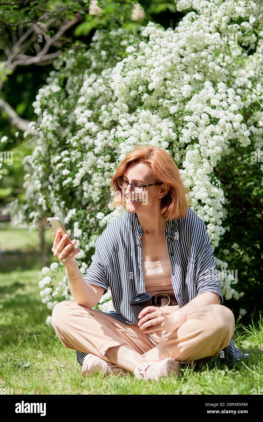Young smiling red-haired woman using her phone, seated outdoors by white flowering blooms with coffee cup. Stock Photo
