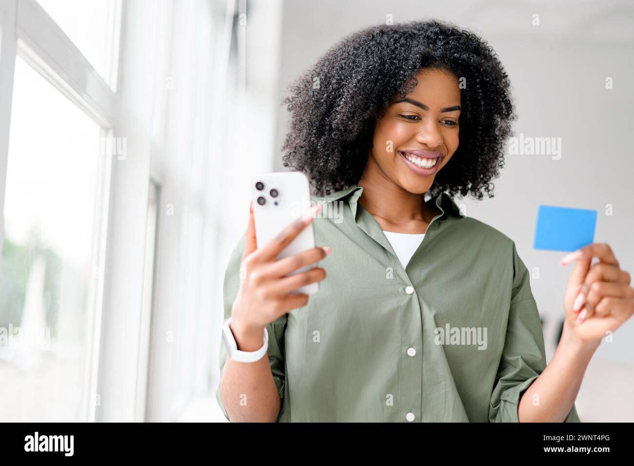 African-American woman beams with pleasure while holding a smartphone and a credit card, possibly making an online purchase or reservation, in a bright, modern interior. Stock Photo