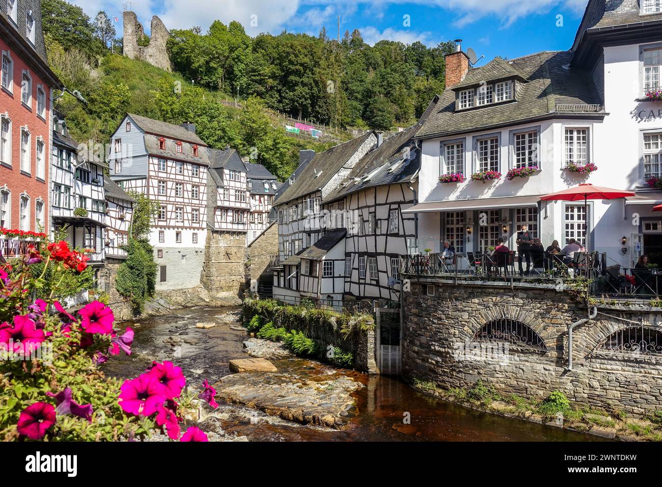 Quaint European village (Monschau, Germany) with half-timbered houses and a flower-lined bridge over a creek, with green hills in the background Stock Photo