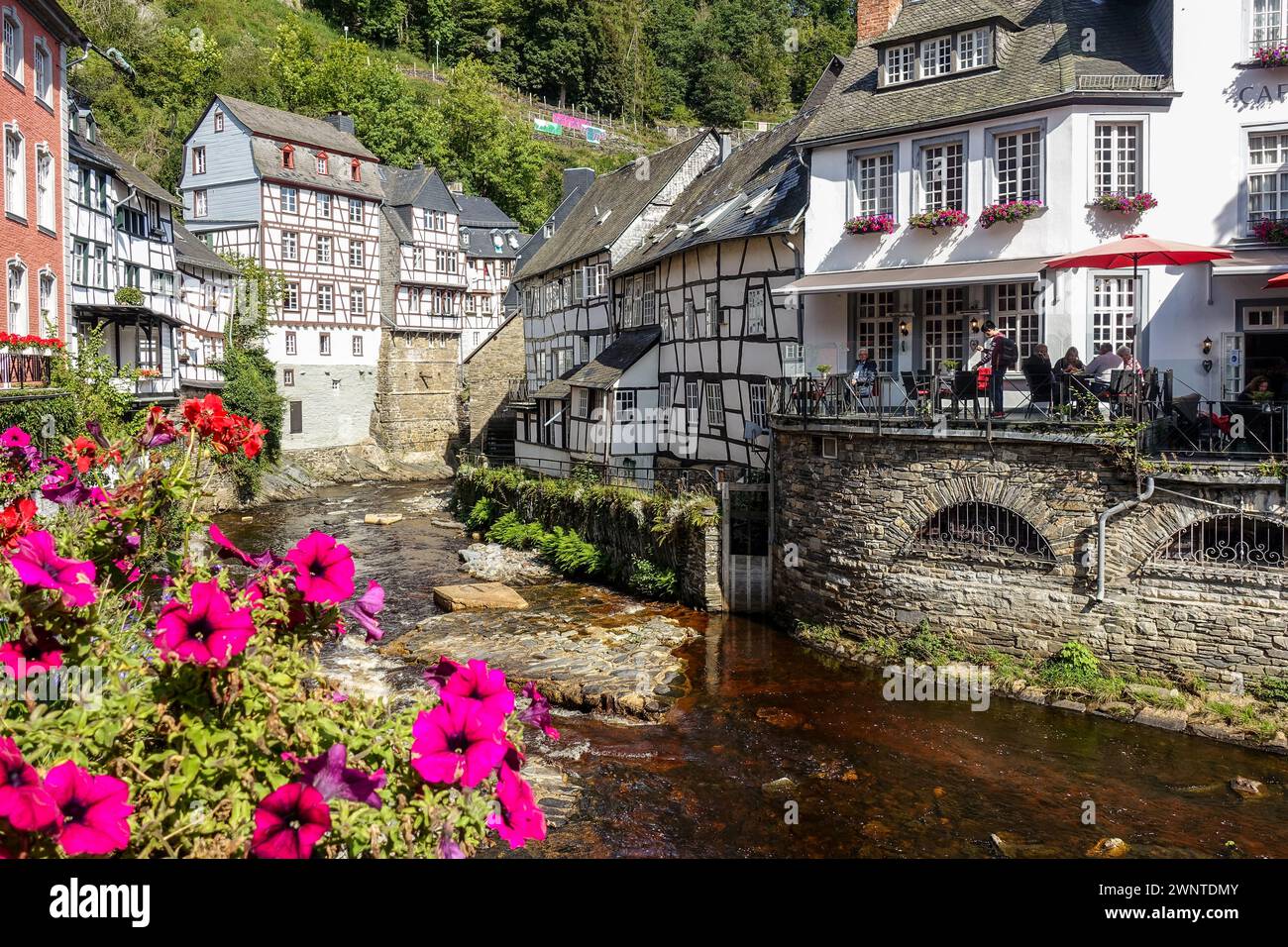 Quaint European village (Monschau, Germany) with half-timbered houses and a flower-lined bridge over a creek, with green hills in the background Stock Photo