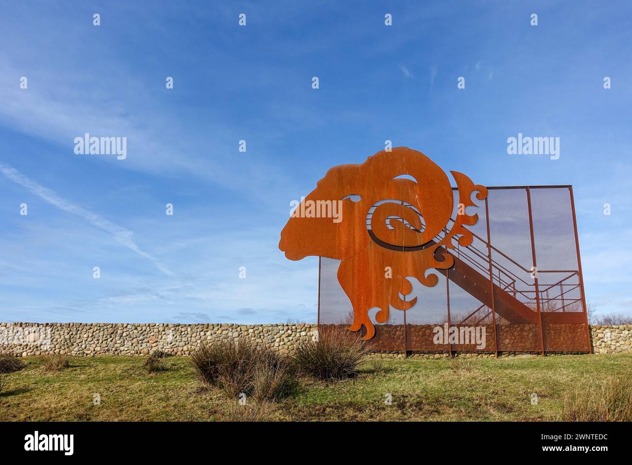 Orange metal ram sculpture in an open field against a blue sky with low stone wall in the background, taken in the Dwingelderveld national park, NL Stock Photo