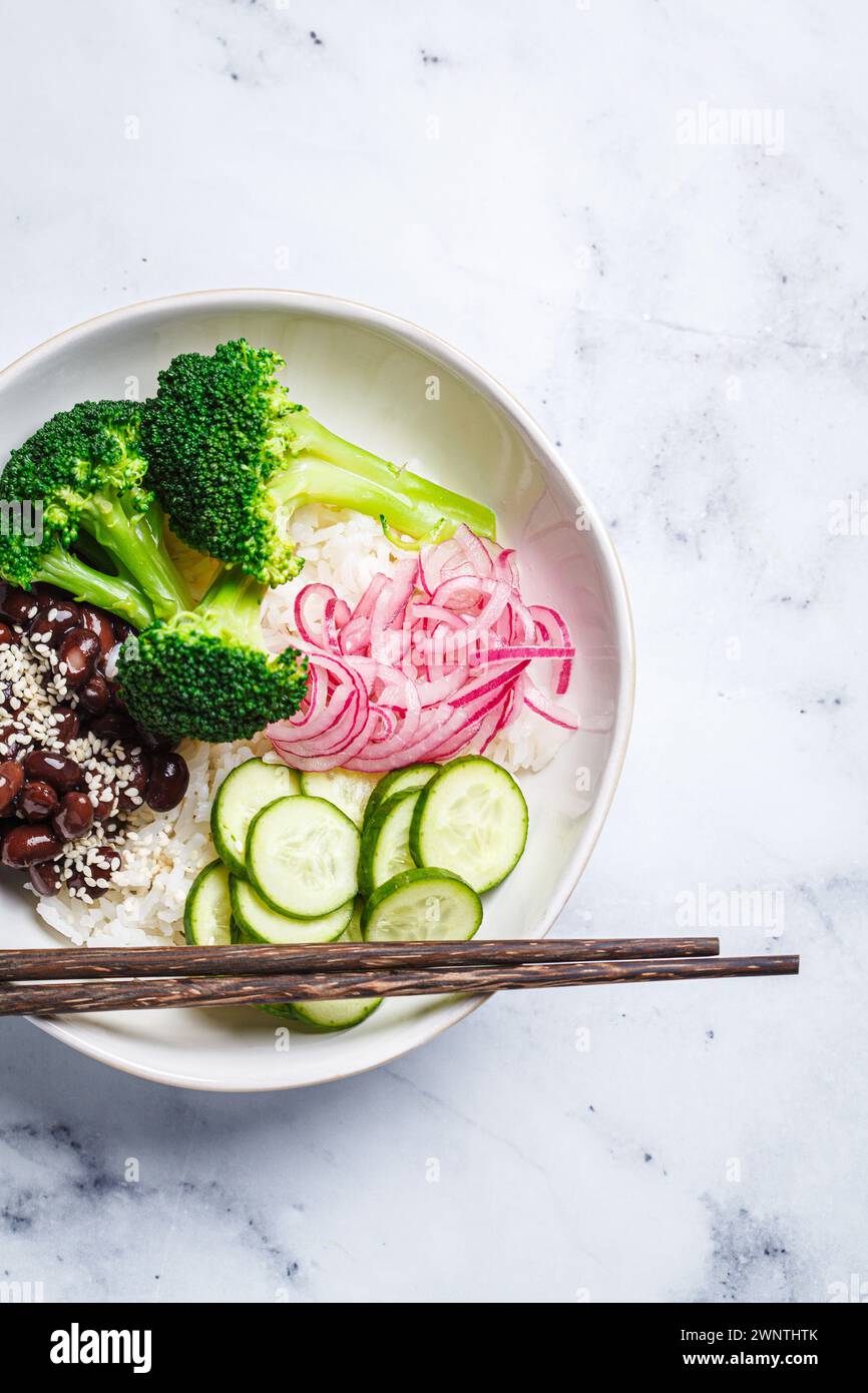 Vegan poke. Bowl with rice, broccoli, cucumber, beans and pickled red onion, white marble background, top view. Stock Photo