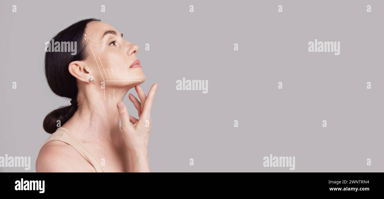 Beautiful senior woman with lifting arrows showing facial anti-aging treatment on skin against grey studio background. Negative space. Stock Photo