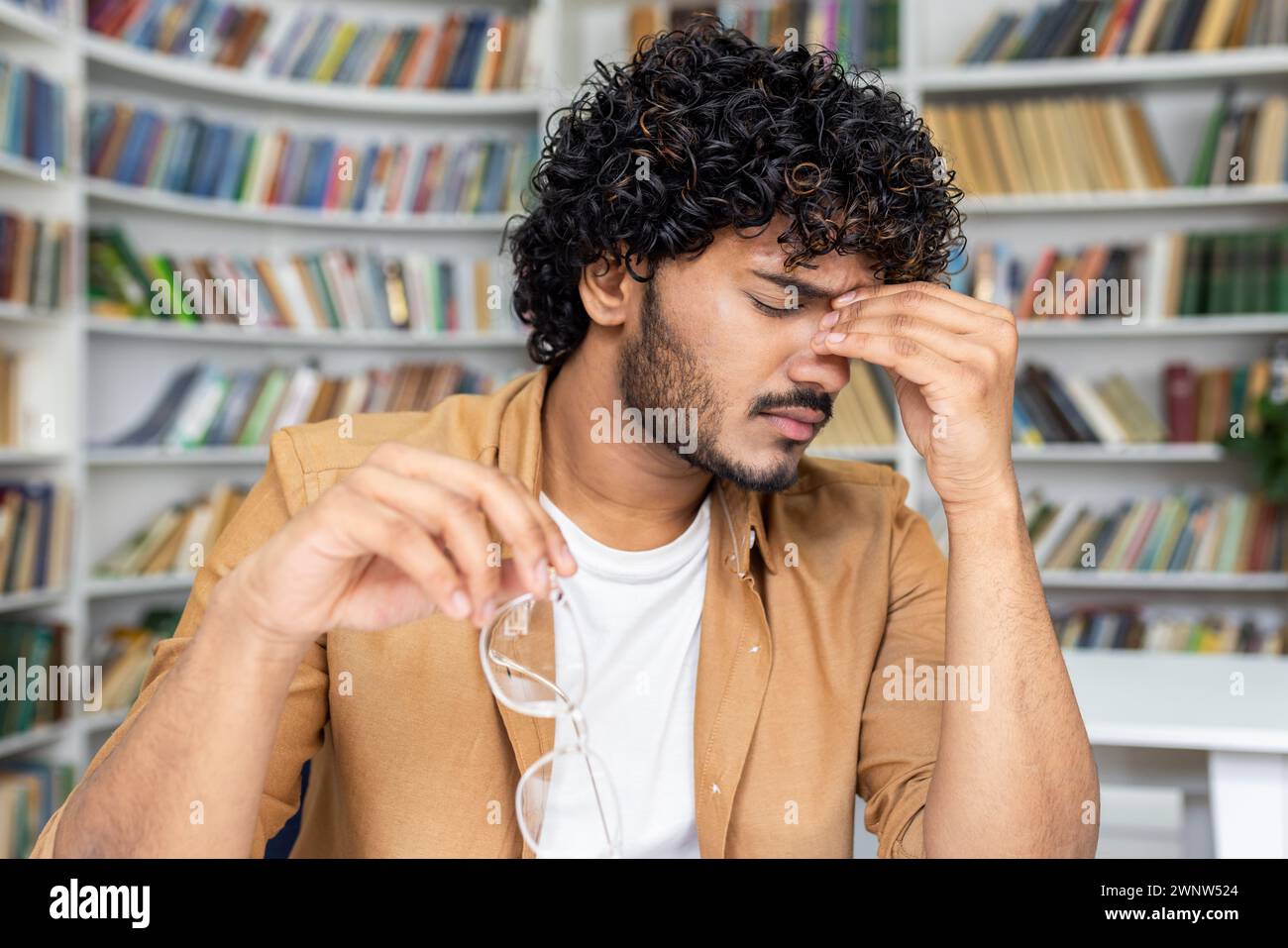 A young man with curly hair appears stressed and holds his head in pain, showing signs of a headache. He has taken off his eyeglasses, perhaps after reading or studying for long hours. Stock Photo