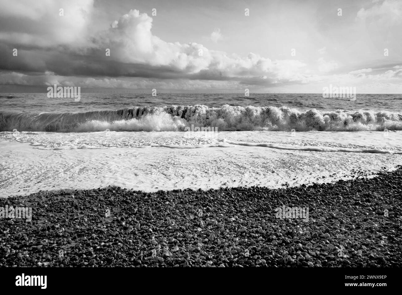 B&w strong sunlight of waves breaking on pebble beach surf, spume & spray, green sea, a blue sky with billowing white clouds South Coast England Stock Photo