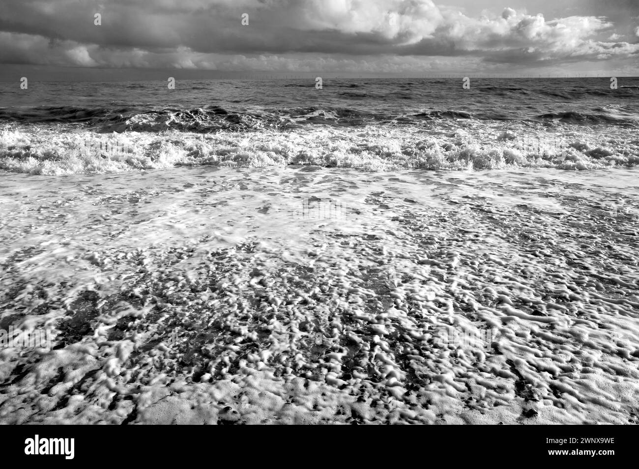 B&w strong sunlight of waves breaking on pebble beach surf, spume & spray, green sea, a blue sky with billowing white clouds South Coast England Stock Photo