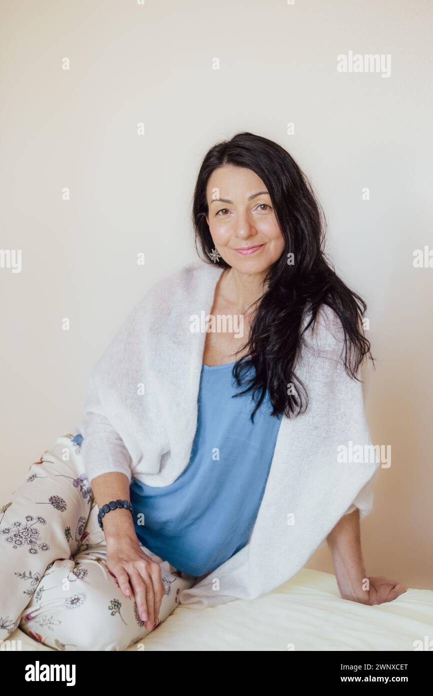 Smiling woman with black hair, blue top, white shawl, sitting comfortably. Her relaxed posture and warm expression evoke a sense of homey contentment. Stock Photo
