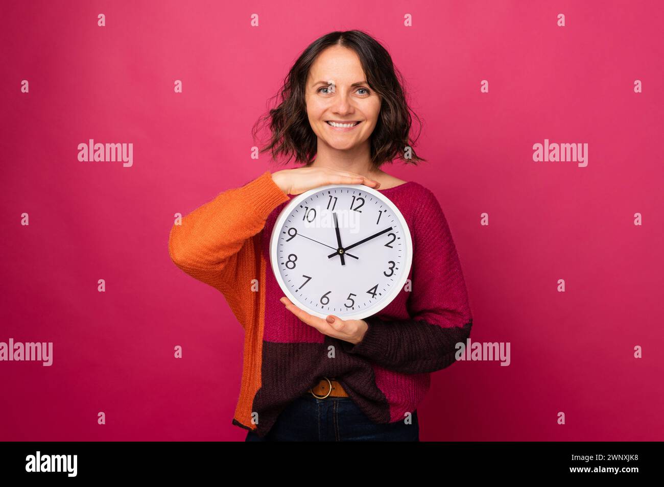 Studio portrait of a beautiful smiling middle age woman holding a round clock over pink background. Stock Photo