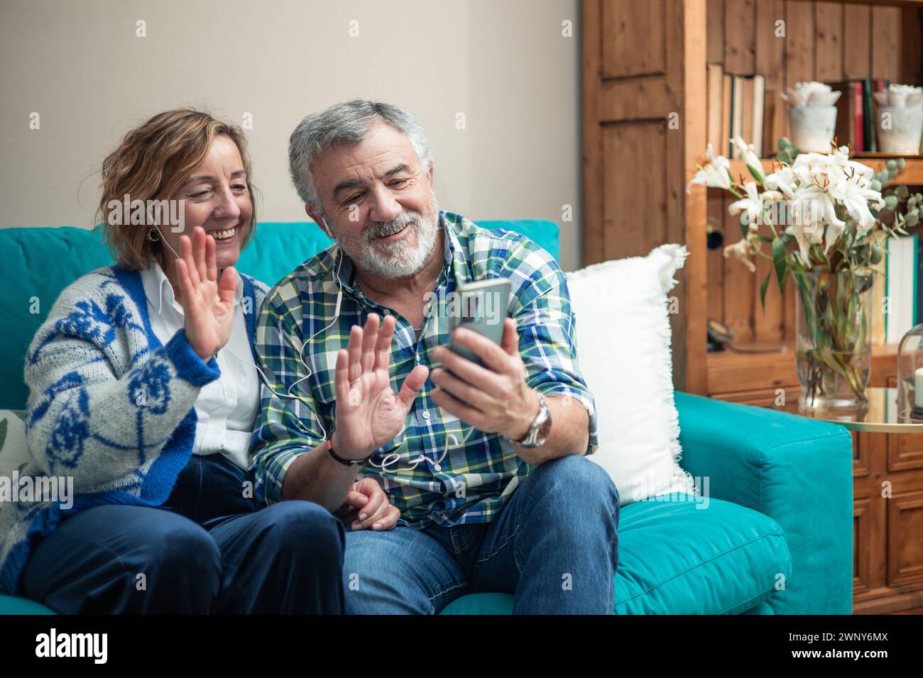 In this modern era, a retired couple sits comfortably on their sofa, warmly greeting their smartphone screen as they engage in a heartwarming video ca Stock Photo