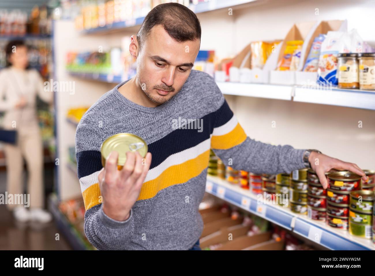 Man chooses canned fish in grocery store. Man shopper reading expiration date on product label