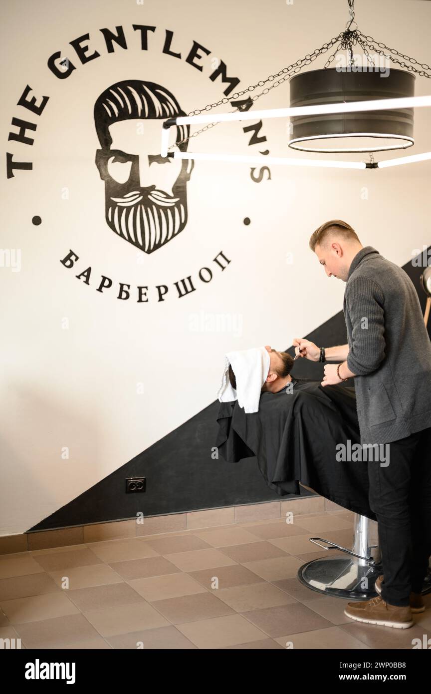 A barber stylist trims the beard of a Caucasian man, whose face is covered with a towel, with scissors Stock Photo