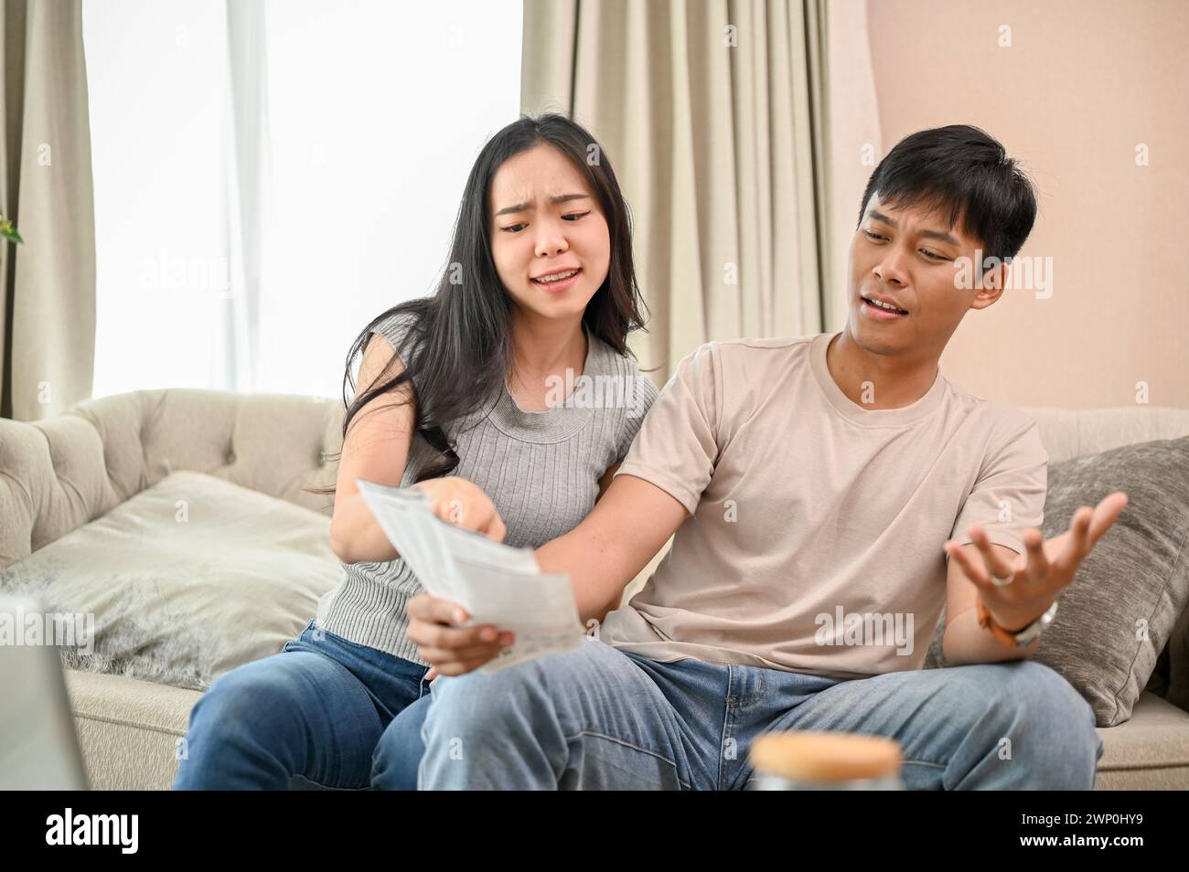 A resentful, angry Asian wife and husband are arguing about family household invoices and high domestic bills, quarreling about the issue of wasting m Stock Photo