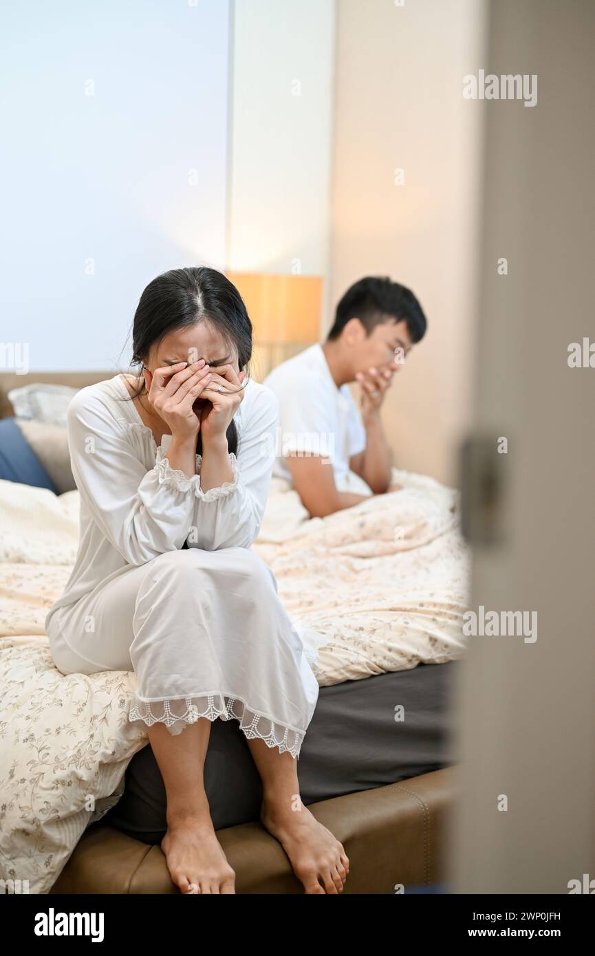 An unhappy, sad Asian wife in pajamas is sitting on the bed, covering her face and crying after an argument with her husband. marriage problems, break Stock Photo