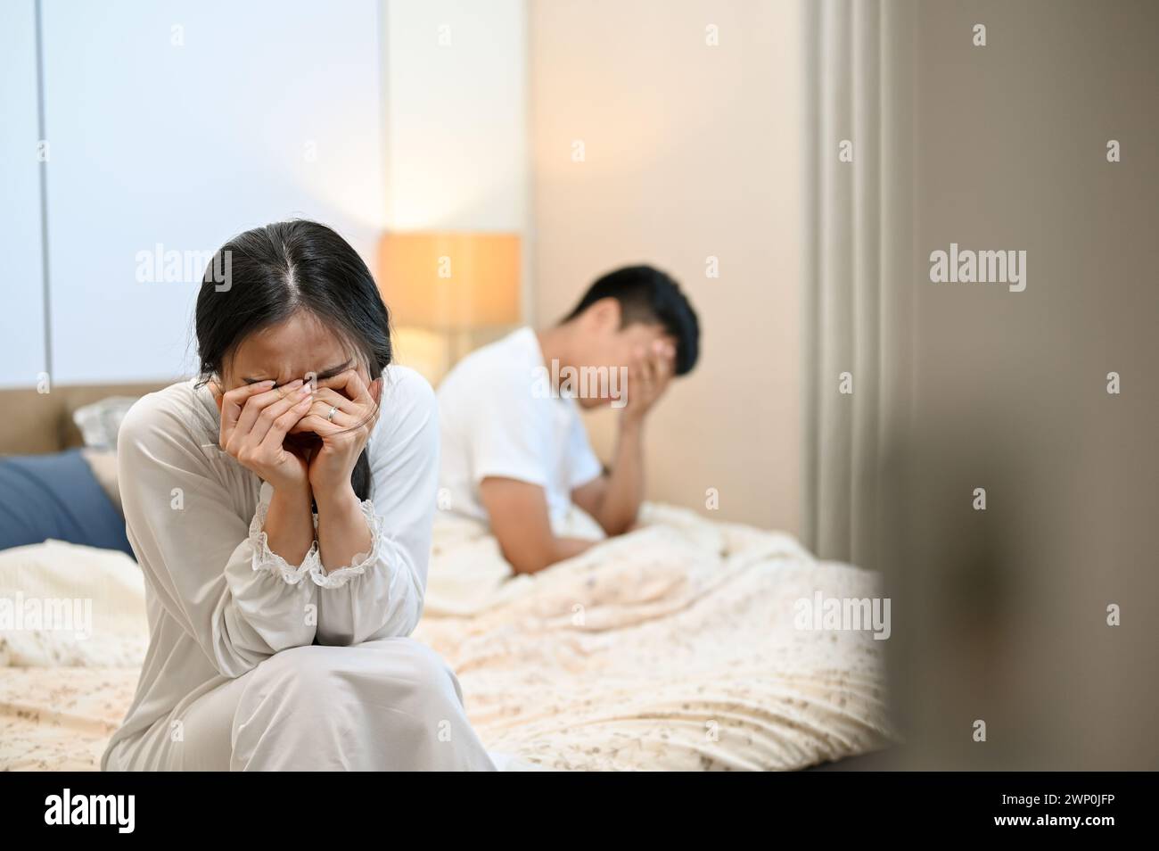 An unhappy, sad Asian wife in pajamas is sitting on the bed, covering her face and crying after an argument with her husband. marriage problems, break Stock Photo