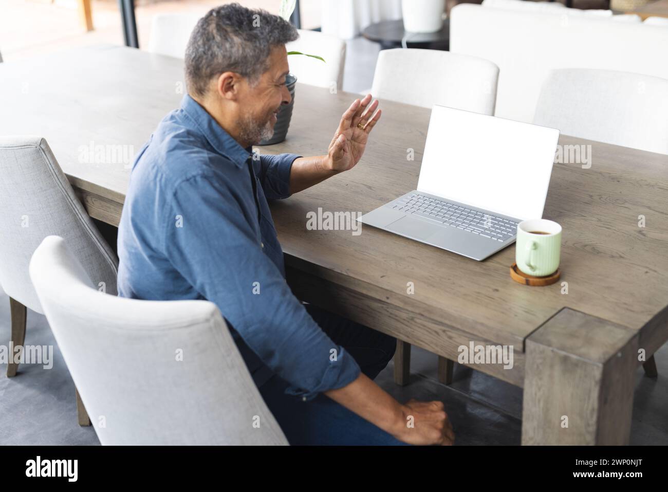 Biracial senior man in a blue shirt waves during a video call on his laptop Stock Photo