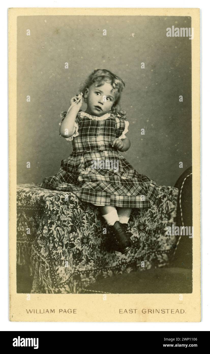 Original, charming Victorian Carte de Visite (visiting card or CDV) of sweet Victorian child with curly hair, wearing button up boots, a beautiful tartan dress, holding up a pendant perhaps with a rattle to it as she appears to be listening to a sound, boys also wore similar dresses in this period. From the studio of  William Page, Moat House, East Grinstead, West Sussex, U.K. circa 1864 Stock Photo