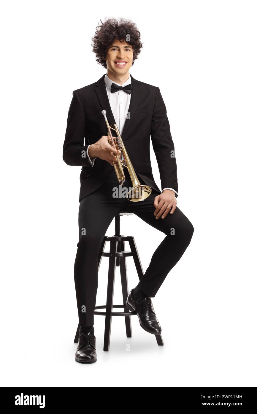 Smiling young male musician in a black suit holding a trumpet and sitting on a chair isolated on white background Stock Photo