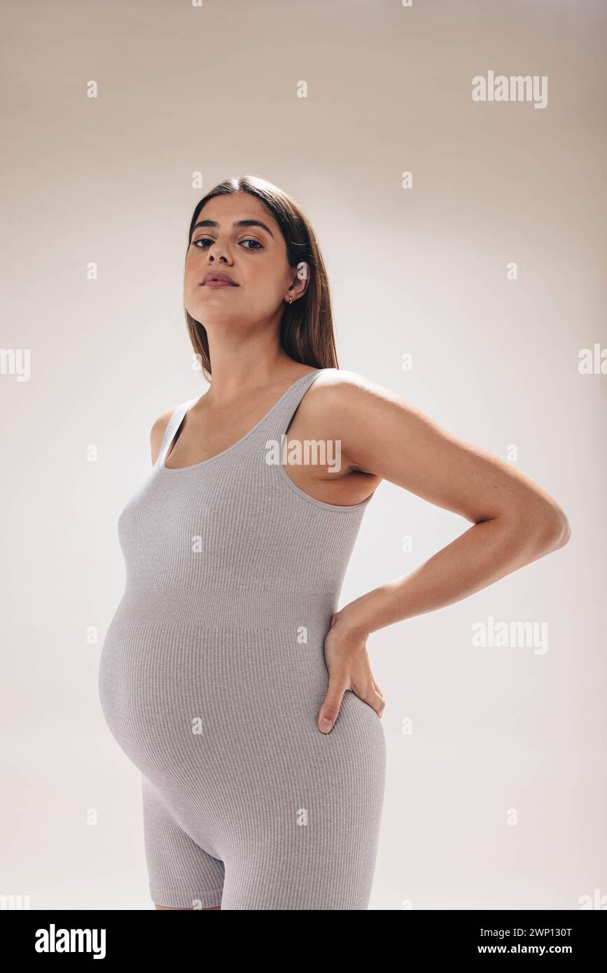 Expecting mother in her third trimester showcases her baby bump while practicing prenatal care in a studio. She wears fitness clothing, exercising for Stock Photo