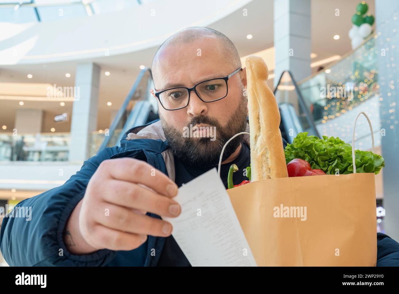 Disappointed plus size man with shopping paper bag full of food looks at sale paper receipt on escalator in supermarket. Male shocked by food Stock Photo