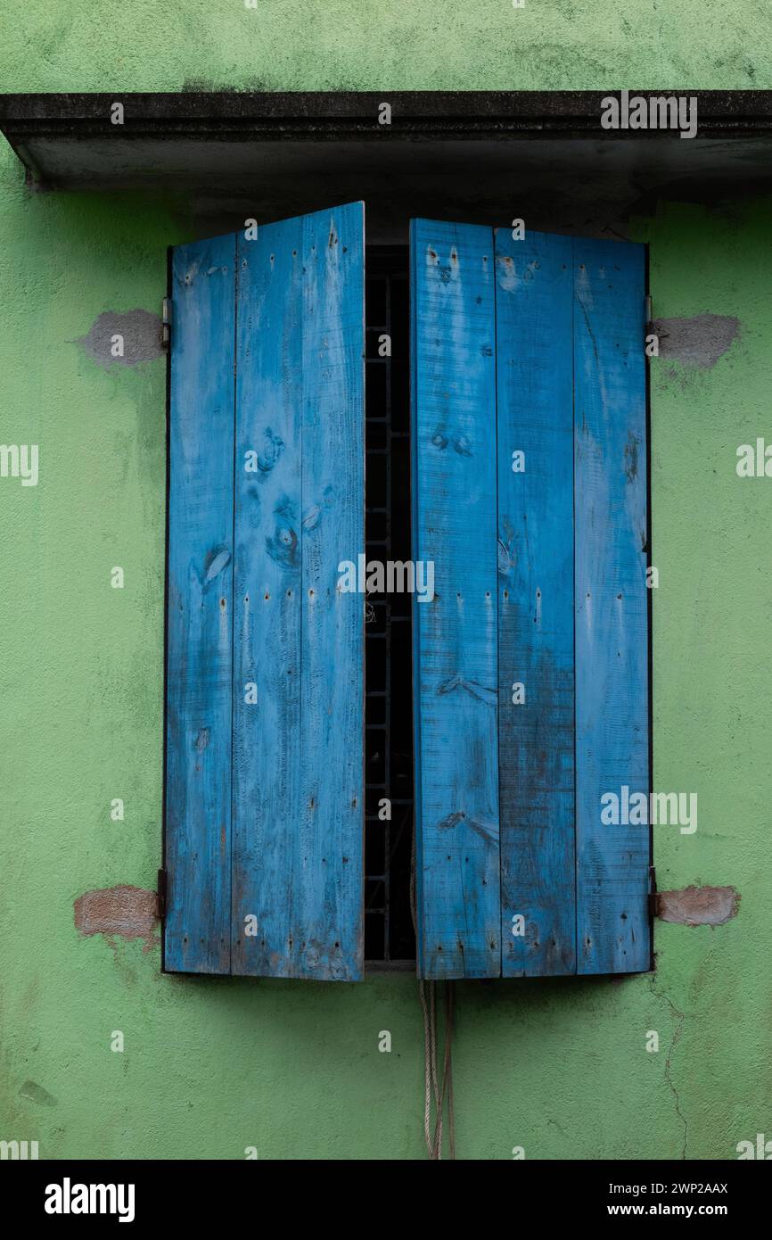 Pair of faded, chipped, painted, slightly open blue wooden shutters against a green wall on a home in rural northern Vietnam. Stock Photo