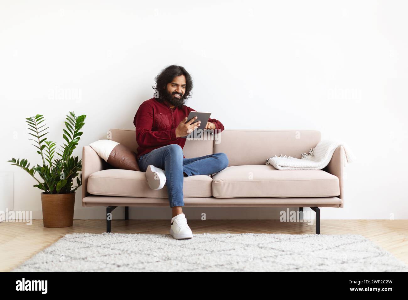 Cheerful young indian man sitting on couch, using digital tablet Stock Photo