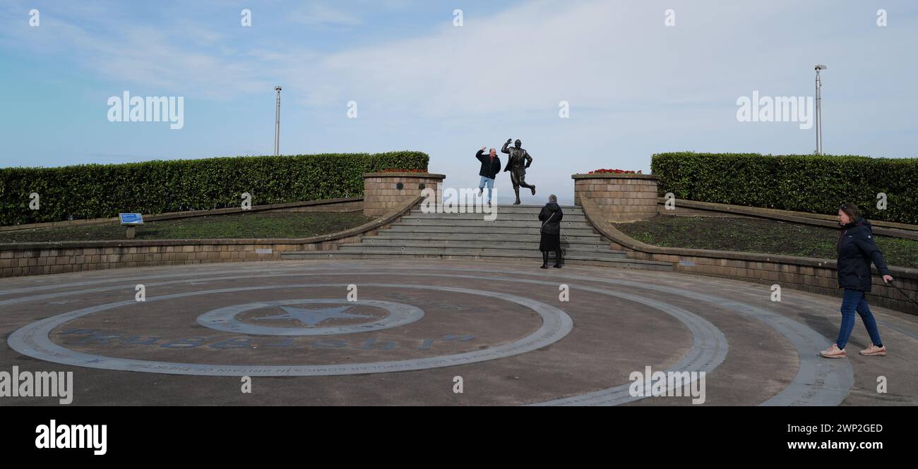 A pan poses for a photograph alongside Eric Morecambe’s statue in Morecambe, Lancashire. Unveiled by the Queen in 1999, the slightly larger than life-sized statue depicts Eric Morecambe in one of his characteristic poses with a pair of binoculars around his neck (he was a keen ornithologist). The statue is set against the stunning backdrop of Morecambe Bay and the Lake District hills Stock Photo