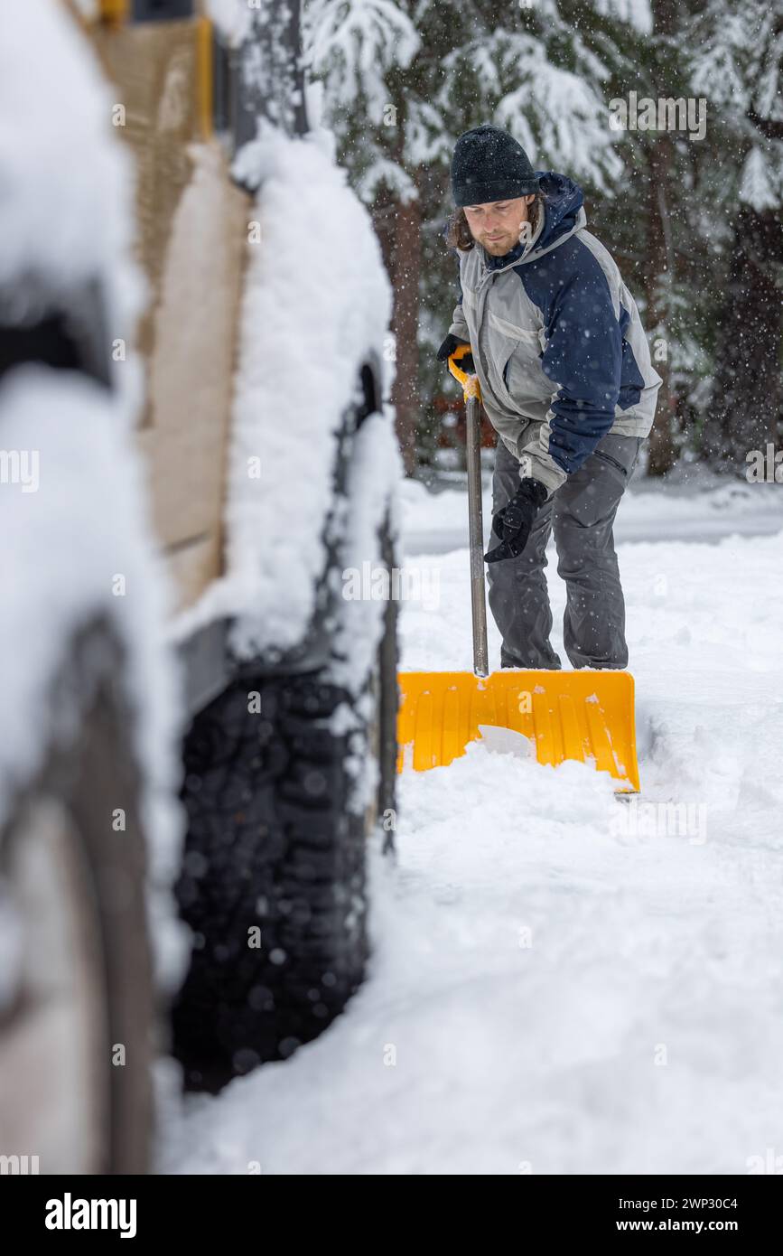 A man is shoveling snow off the ground. He is wearing a black hat and a blue jacket Stock Photo