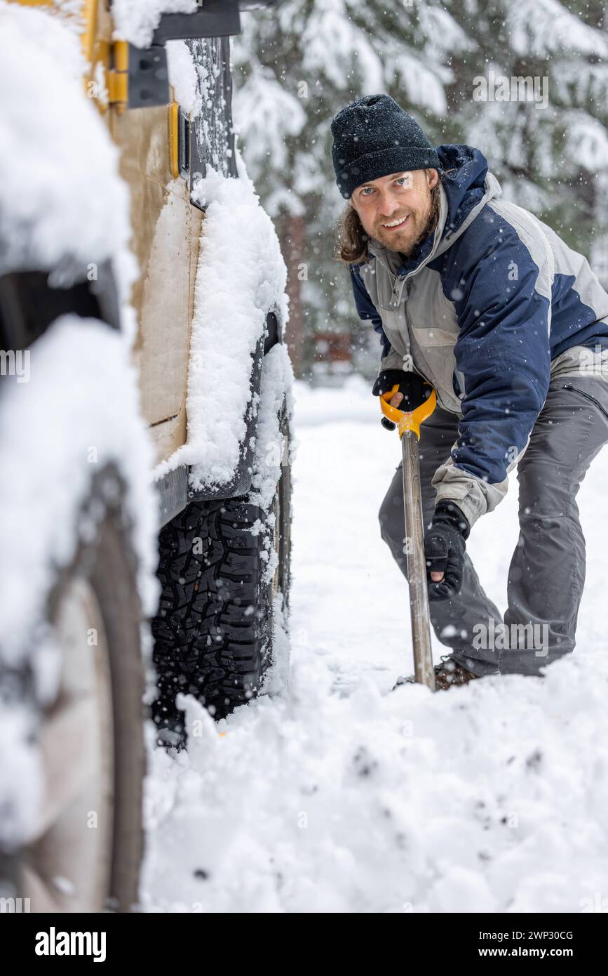 A man in a blue jacket and hat is shoveling snow off the back of a truck. The man is smiling and he is enjoying the task Stock Photo