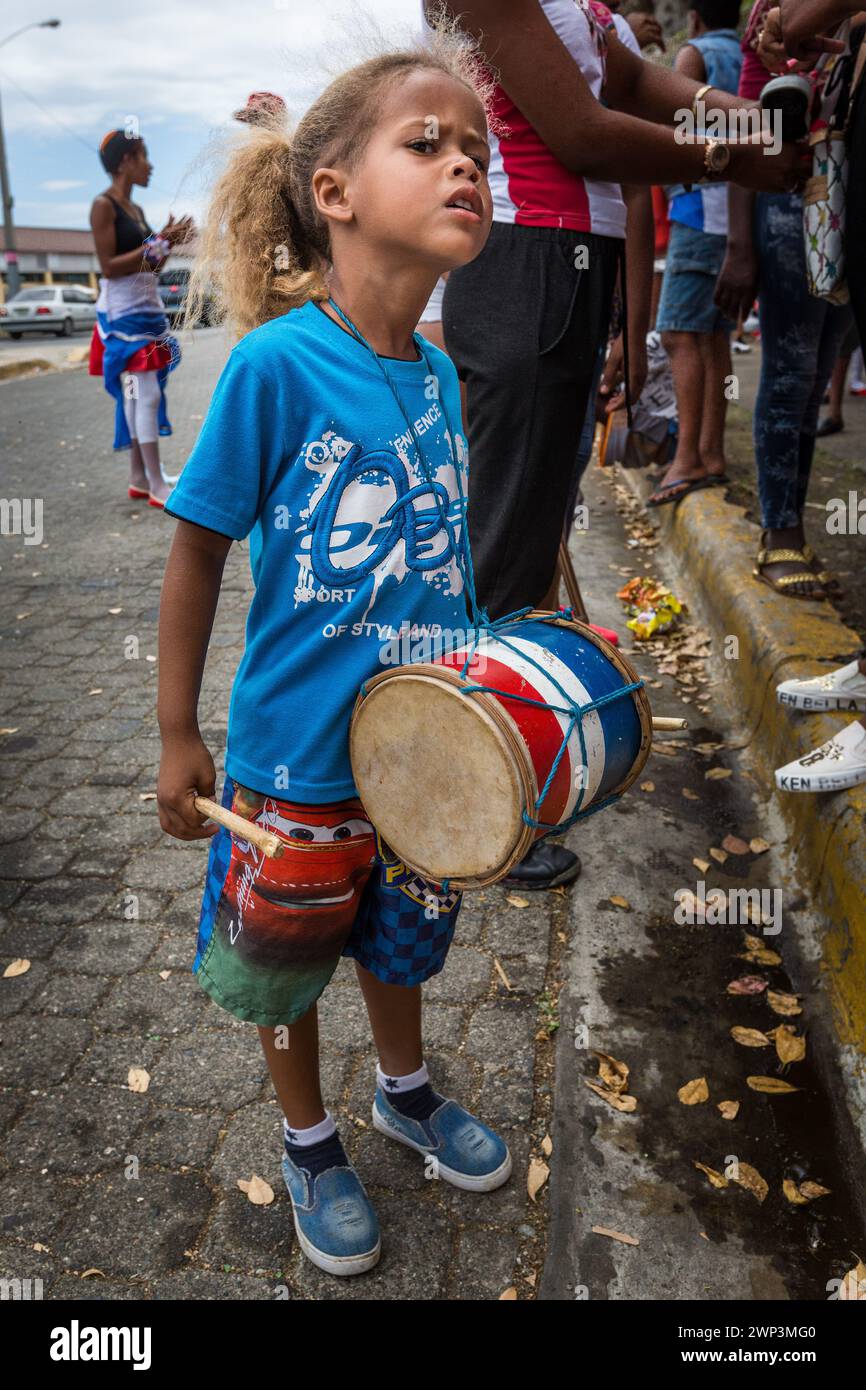 A little Dominican girl with her tambura drum waits for the youth Carnival parade in Santo Domingo, Dominican Republic.  UNESCO World Heritage Site - Stock Photo