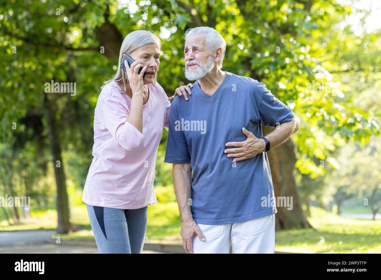 An elderly couple walks in the park, a gray-faced man stands and holds his hand to his chest, feels pain in his heart, a woman supports him and calls for emergency help. Stock Photo