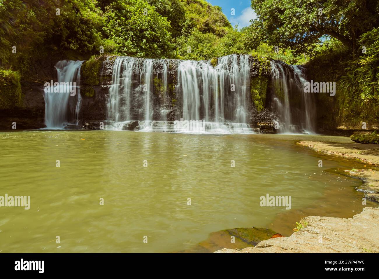 Landscape of a waterfall taken with ND filter with trees and blue sky in the background and water basin in the foreground in Guam Stock Photo