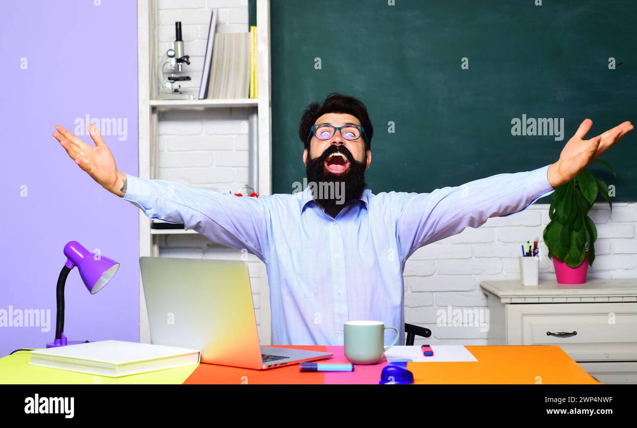 Portrait of happy male teacher or student in university. Bearded man professor at school lesson in classroom using laptop sitting at desk. Virtual Stock Photo