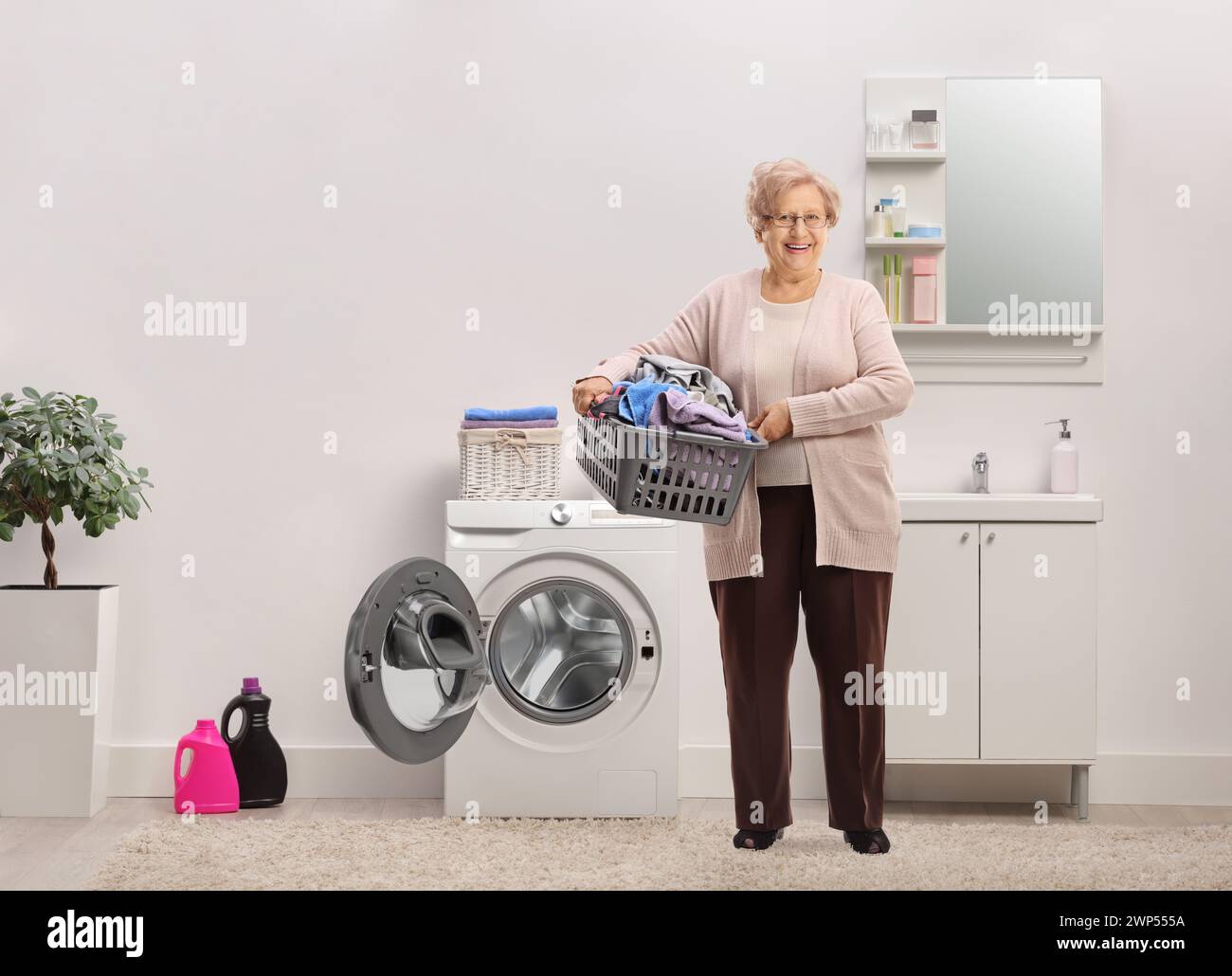 Full length portrait of an elderly lady holding a laundry basket with clothes inside a bathroom Stock Photo