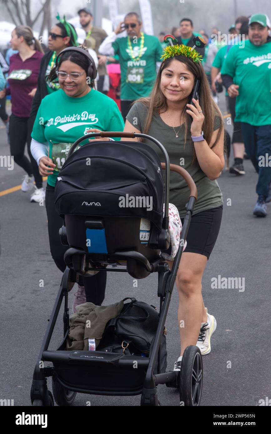 Crowd of people in green t-shirts, a woman on a cell phone pushing a baby carriage, during St. Patrick's Day Run & Walk, Pharr, Texas, USA. Stock Photo