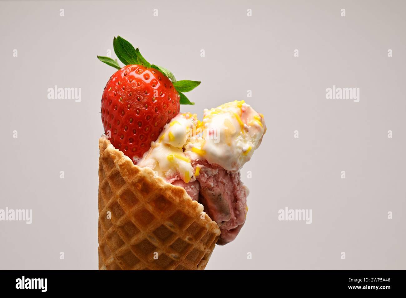 Delicious vanilla and berry ice cream scoops in a waffle cone topped with a fresh strawberry and mint leaf against a white background Stock Photo