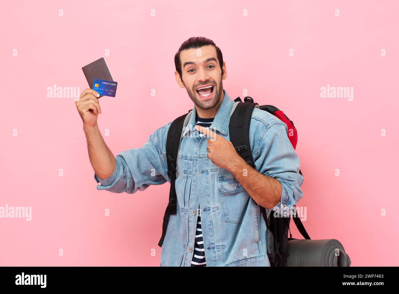 Smiling solo tourist man backpacker pointing to passport and credit card in his hand on pink color studio isolated background Stock Photo