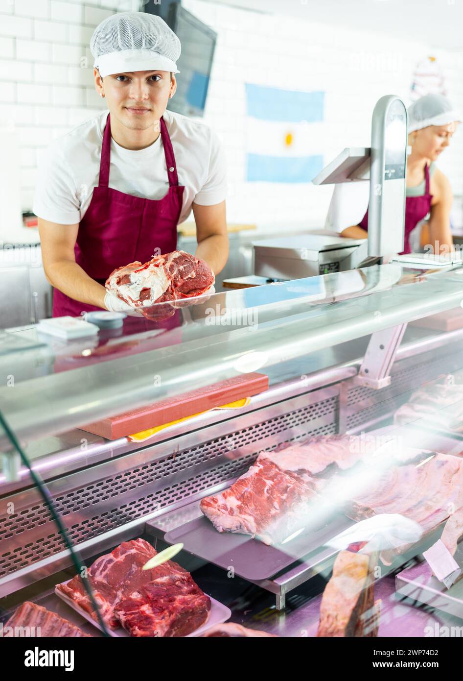 Positive young salesman demonstrating piece of meat in butcher shop ...