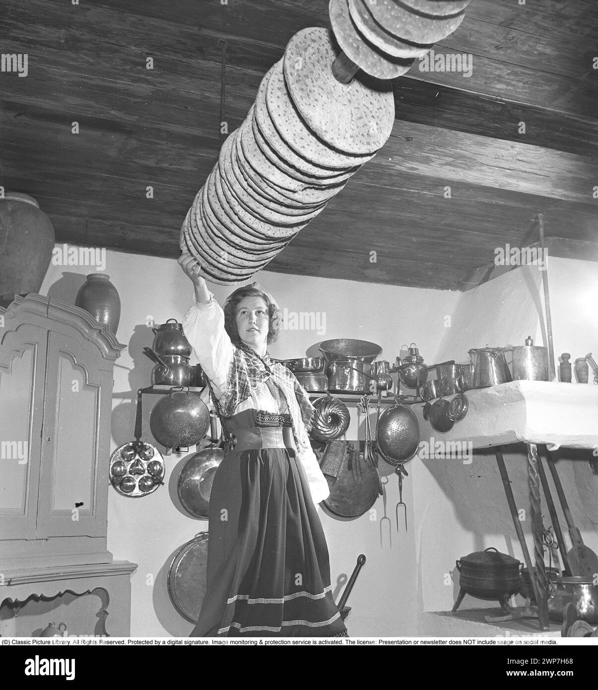 Baking bread 1950. Interior of a kitchen with a women dressed in a traditional swedish  handling crispbread. A flat and dry type of bread, containing mostly rye flour. The baking oven is heated with firewood.  The cripsbread is stored on a rack hanging from a pole in the ceiling, thanks to the hole made in the middle of the cripsbread. A rural kitchen with a wooden fireplace and historical objects as decoration. Sweden 1950. Kristoffersson ref AY55-3 Stock Photo
