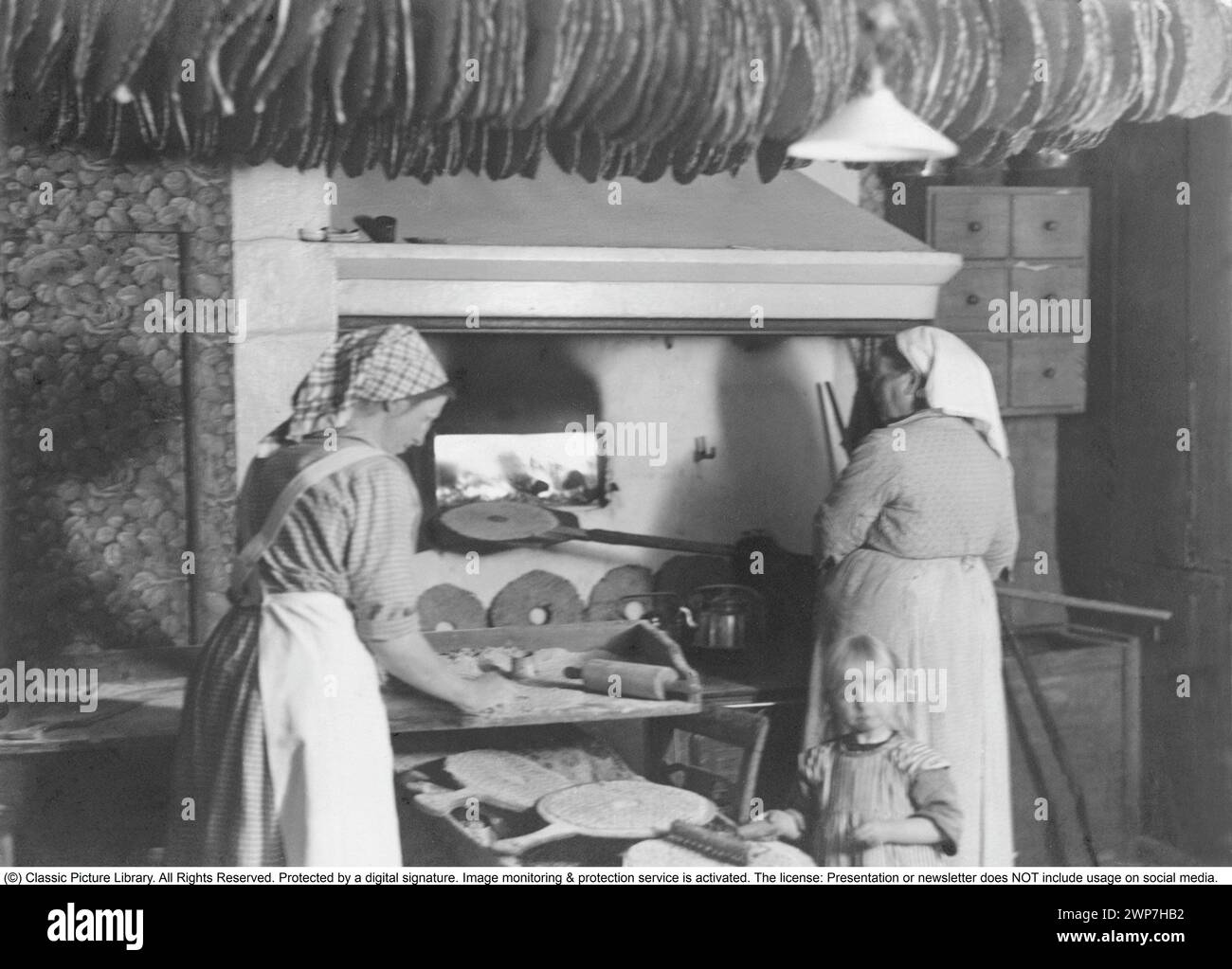 Baking bread 1920s. Interior of a kitchen with women baking crispbread. A flat and dry type of bread, containing mostly rye flour. The baking oven is heated with firewood.  The traditional baking tool like rolling pins is visible and a rack of ready baked bread hanging from a pole in the ceiling, thanks to the hole made in the middle of the cripsbread. Picture taken in the small locality Ransäter in Värmland Sweden. Stock Photo