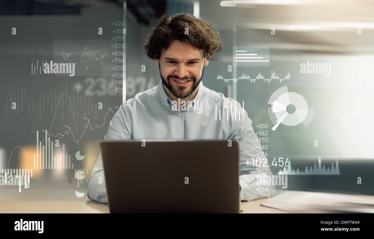 A cheerful bearded man engaging with a laptop in a modern office setting, digital graphs Stock Photo