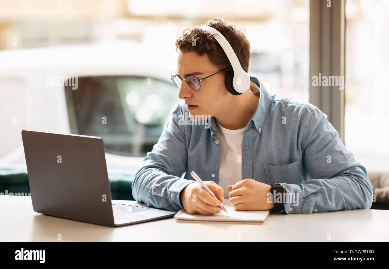 A young, curly-haired man wearing headphones writes in a notebook while working Stock Photo