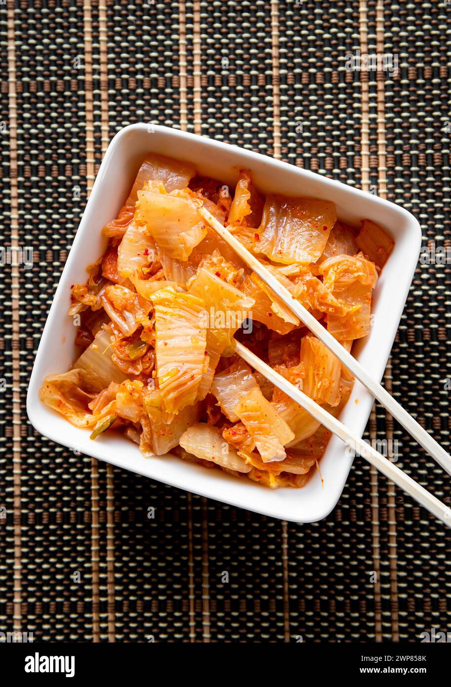 Selective focus on bamboo chopsticks with fermented kimchi cabbage salad and white food bowl on brown background. Stock Photo