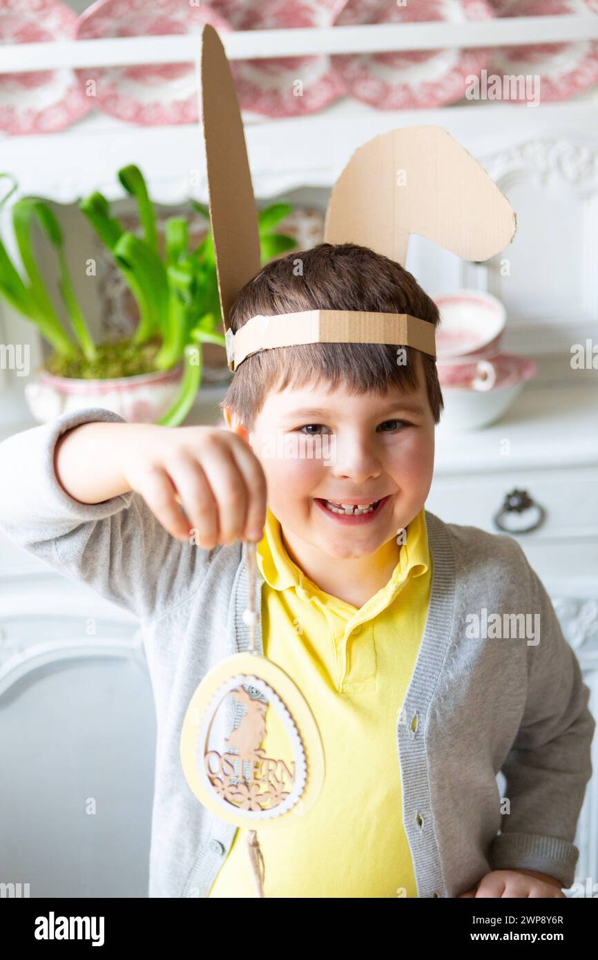 3 March 2024: Little boy with self-made bunny ears holding decoration with Easter written on it. Decorating and crafting for Easter concept *** Kleiner Junge mit selbstgebastelten Hasenohren hält Dekoration mit Schriftzug Ostern in der Hand. Dekorieren und Basteln zu Ostern Konzept Stock Photo