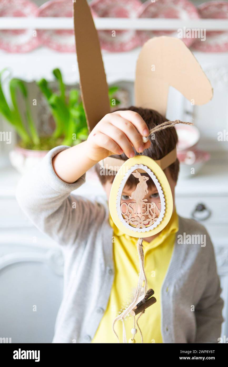 3 March 2024: Little boy with self-made bunny ears holding decoration with Easter written on it. Decorating and crafting for Easter concept *** Kleiner Junge mit selbstgebastelten Hasenohren hält Dekoration mit Schriftzug Ostern in der Hand. Dekorieren und Basteln zu Ostern Konzept Stock Photo