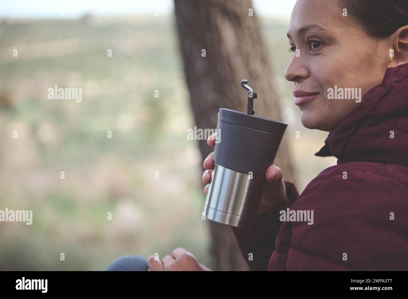 Close-up portrait of a happy smiling woman, traveler, female adventurer, tourist hiker drinking hot tea or coffee from a travel mug while resting afte Stock Photo