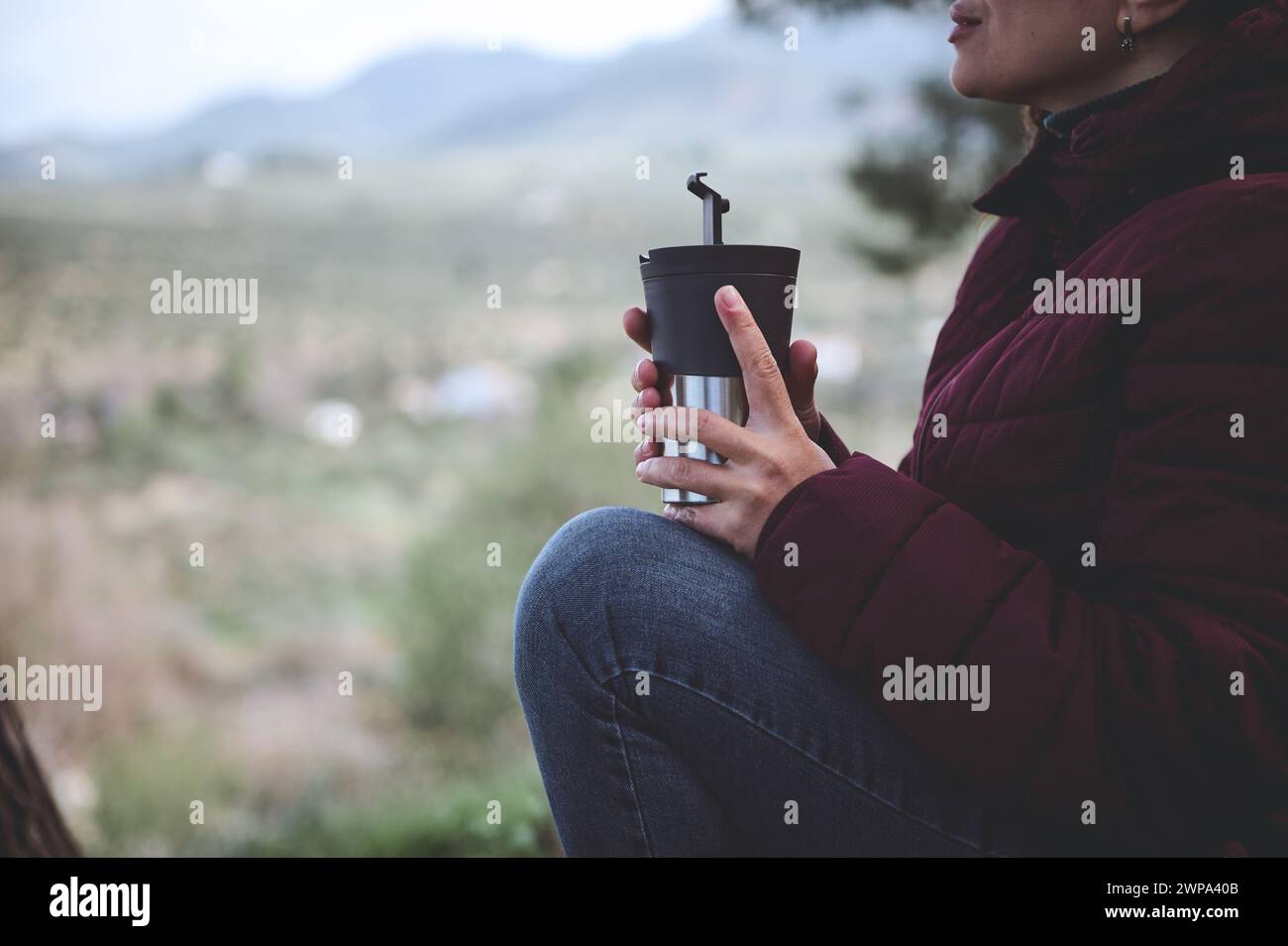 Female traveler in warm winter clothes, holding a thermos mug with hot drink, warming hands while relaxing after hiking in the forest or mountains. Pe Stock Photo
