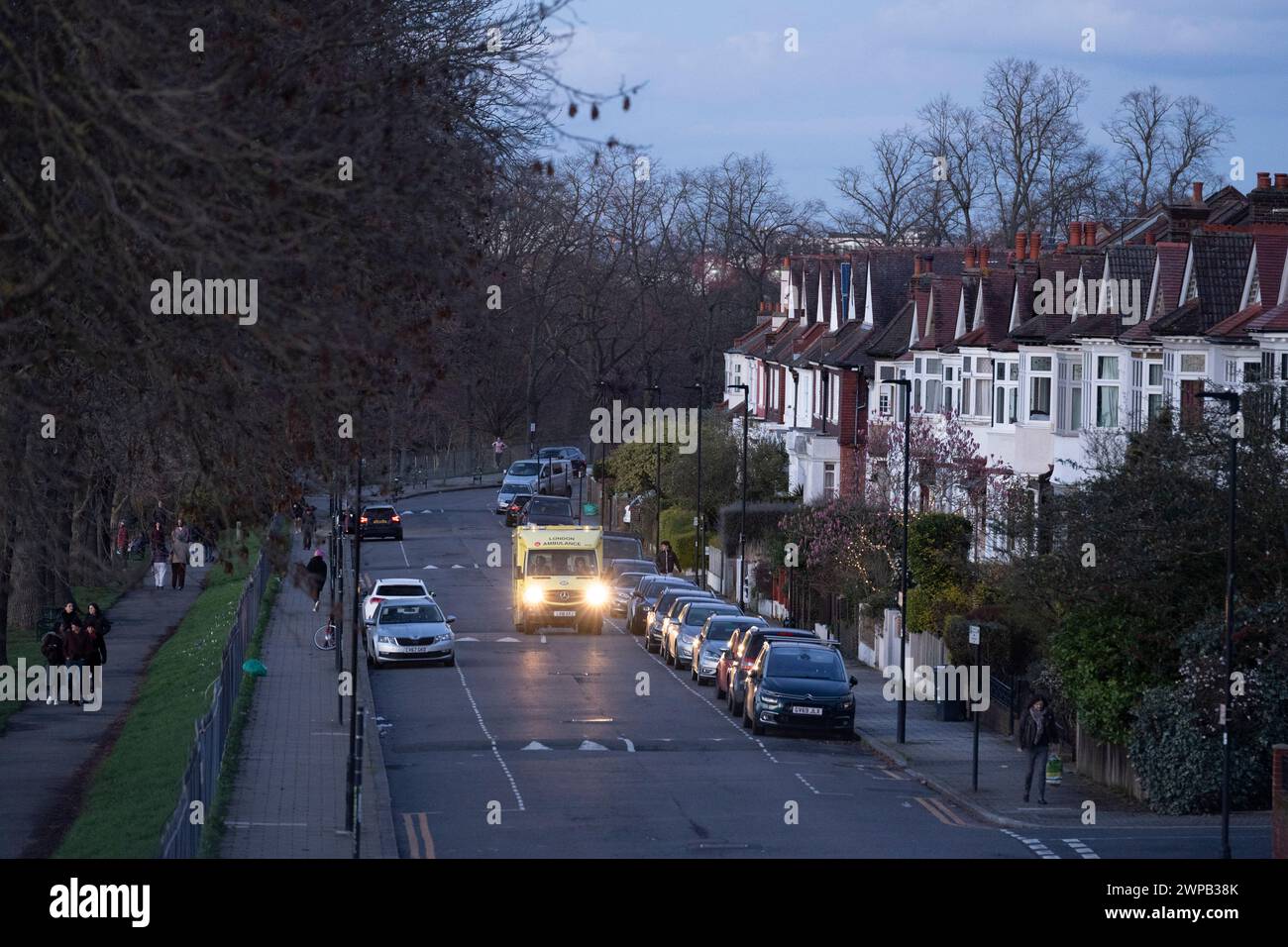 An NHS ambulance drives down a suburban street lined with residential properties, parked cars and park users walking in Ruskin Park in south London, Lambeth, on 2nd March 2024, in London, England. Stock Photo