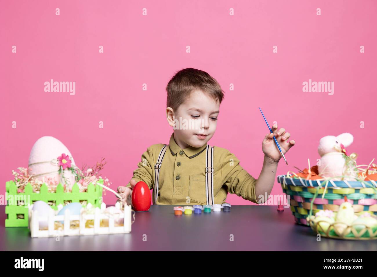 Lovely youngster with a smile crafting eggs and decorations for the Easter festivities, using art supplies and watercolor paint. Little pleased boy feeling excited about painting activity. Stock Photo