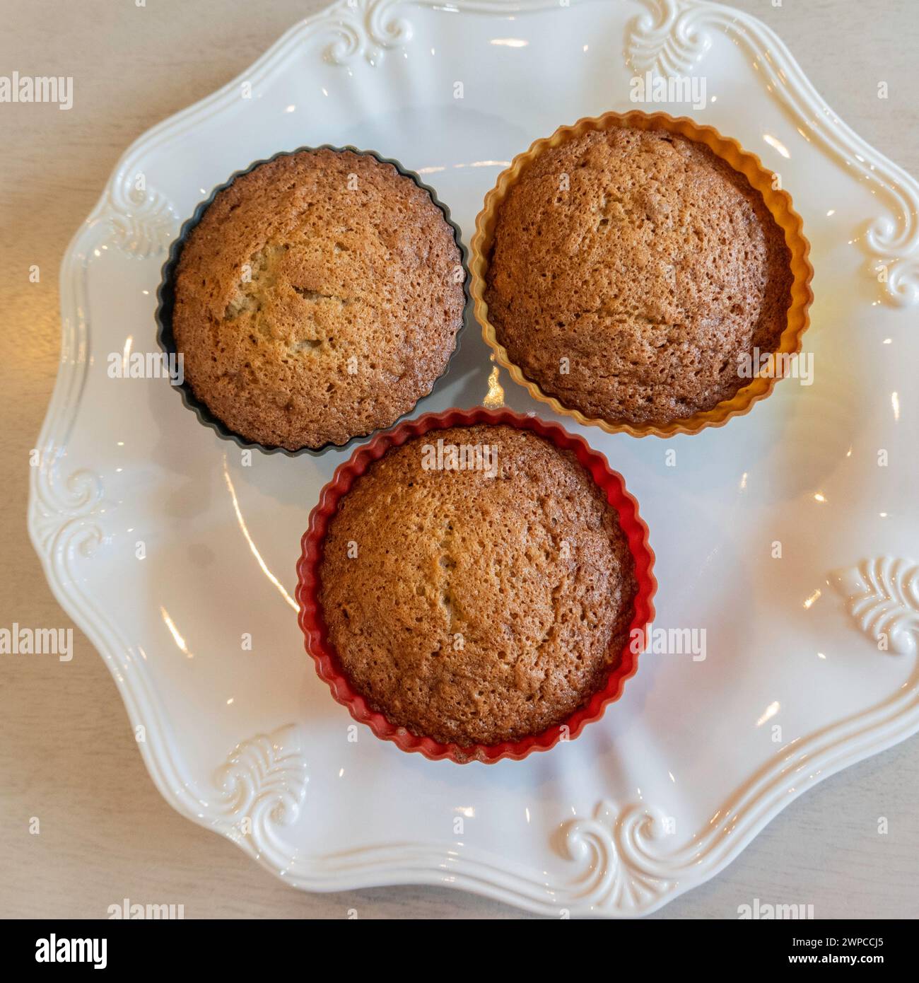 Three homemade fresh-baked banana nut muffins in silicone cups on a white plate taken from above. USA. Stock Photo