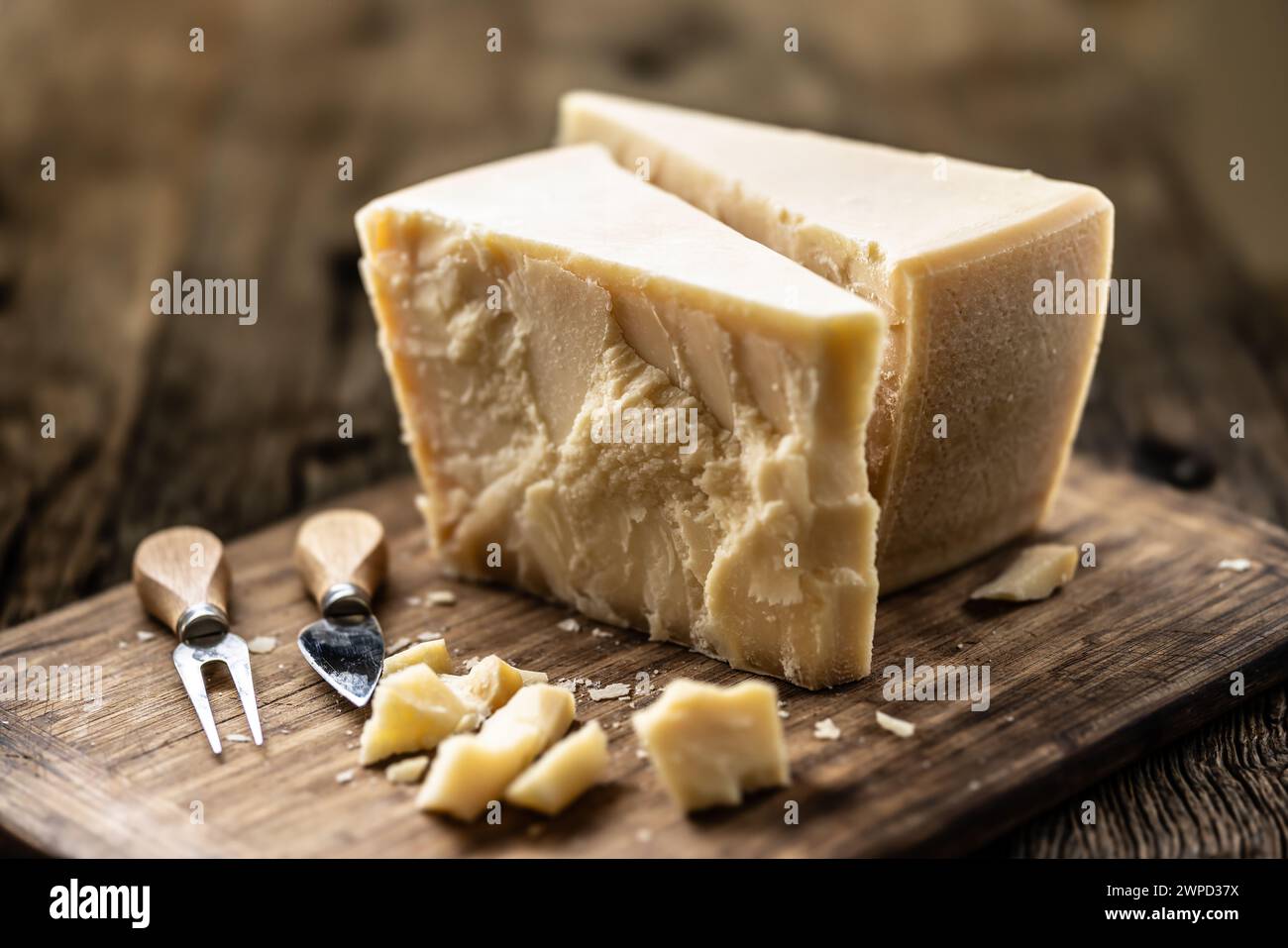 A block and pieces of Parmesan cheese on a cutting board along with a chisel and a skewer. Stock Photo